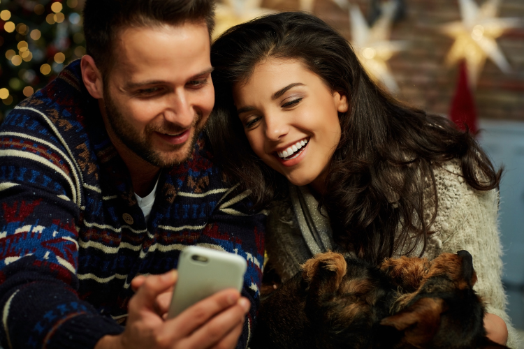 Christmas couple at home in Winter. Happy young couple lying on floor using smart phone at home with dog at Christmas time. Christmas tree and fireplace in background.