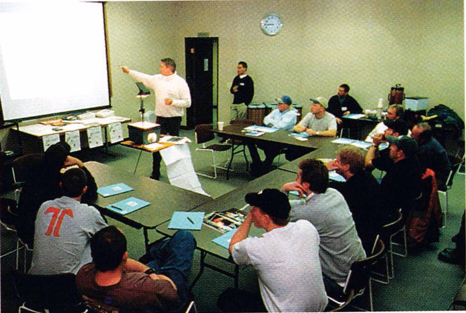 Participants seated at tables in a classroom session while a presenter stands at the front