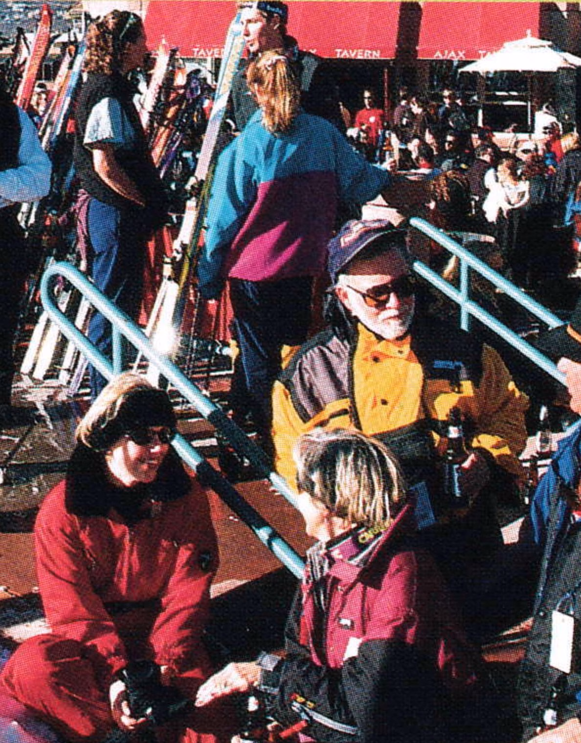 Crowd of skiers relaxing on a sunny outdoor deck, with skis racked behind them