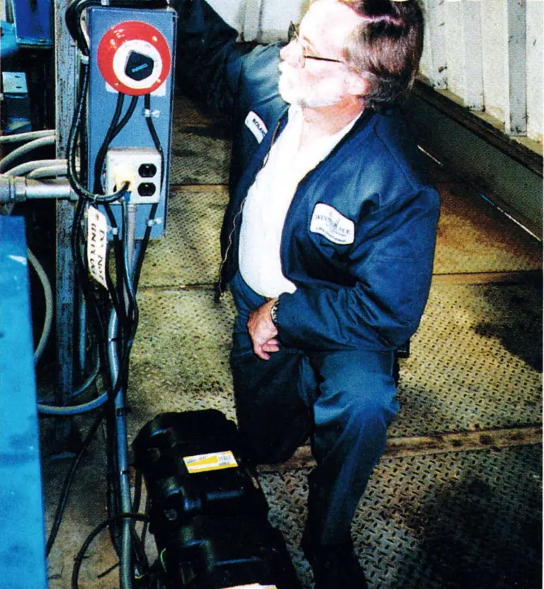 Lift mechanic kneeling beside two auxiliary batteries and a red battery switch in a lift drive room