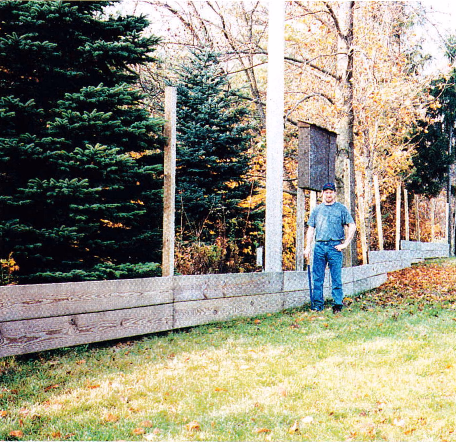 Man standing beside a wooden board barrier holding snow at a hilltop lift unloading area, with trees behind