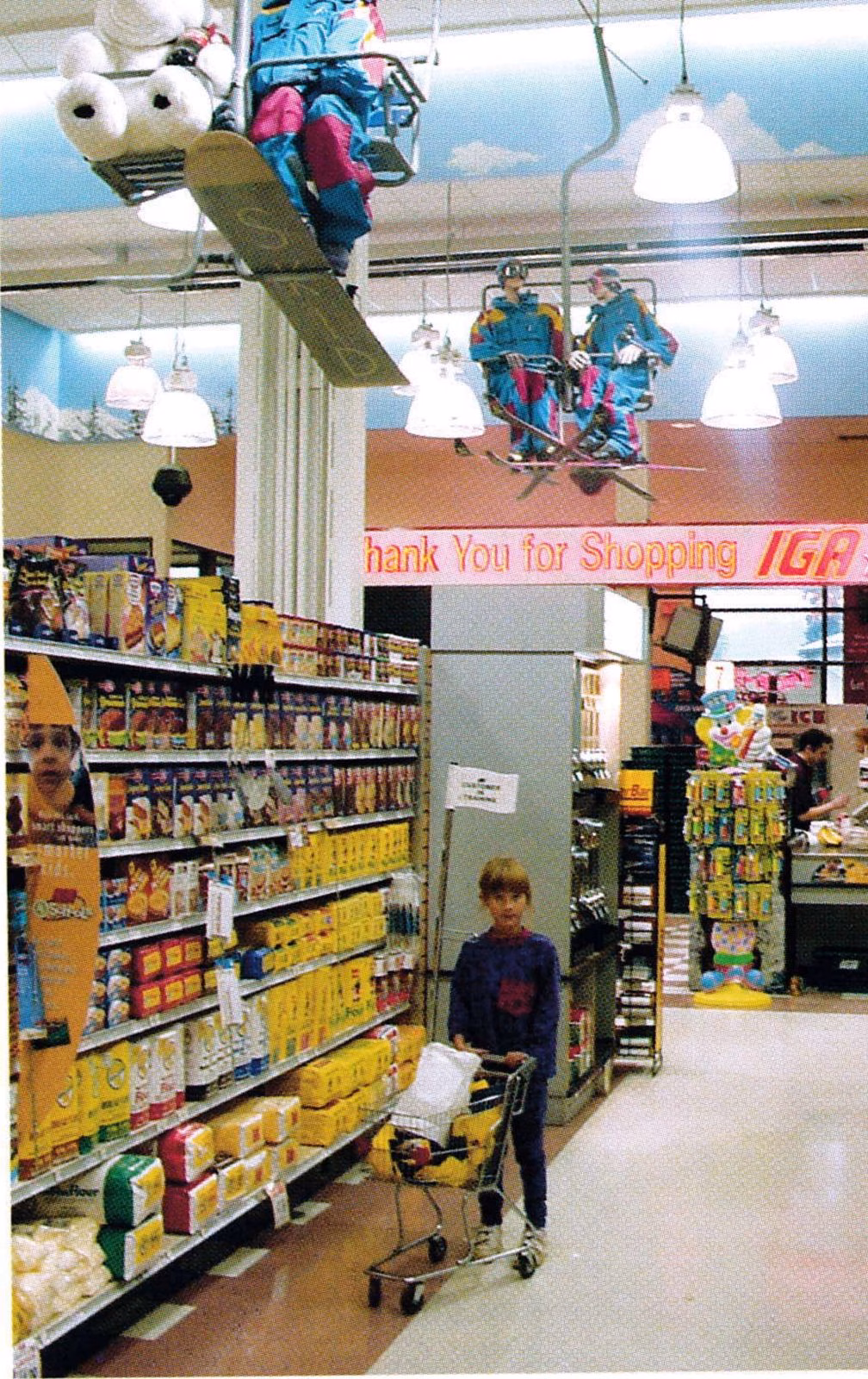 Grocery store aisle with old chairlift chairs holding ski-dressed mannequins hung from the ceiling, a "Thank You for Shopping IGA" sign, and a child pushing a small shopping cart