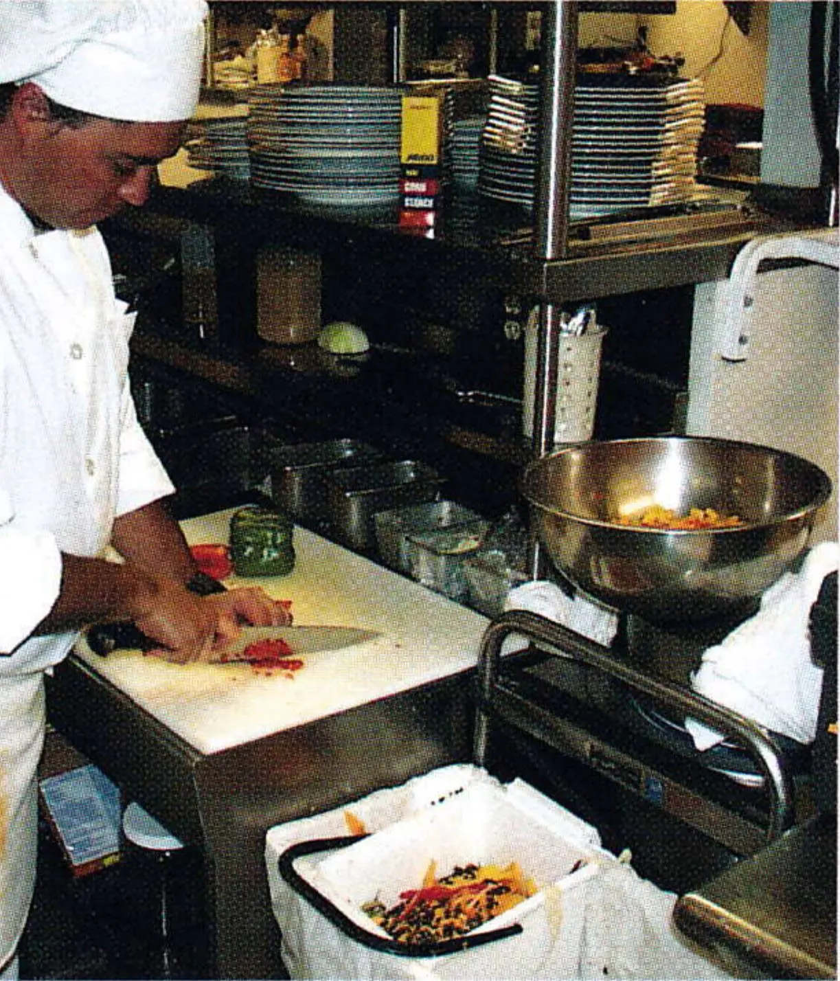 Cook in white uniform cutting vegetables at a restaurant kitchen counter beside a bag of food scraps