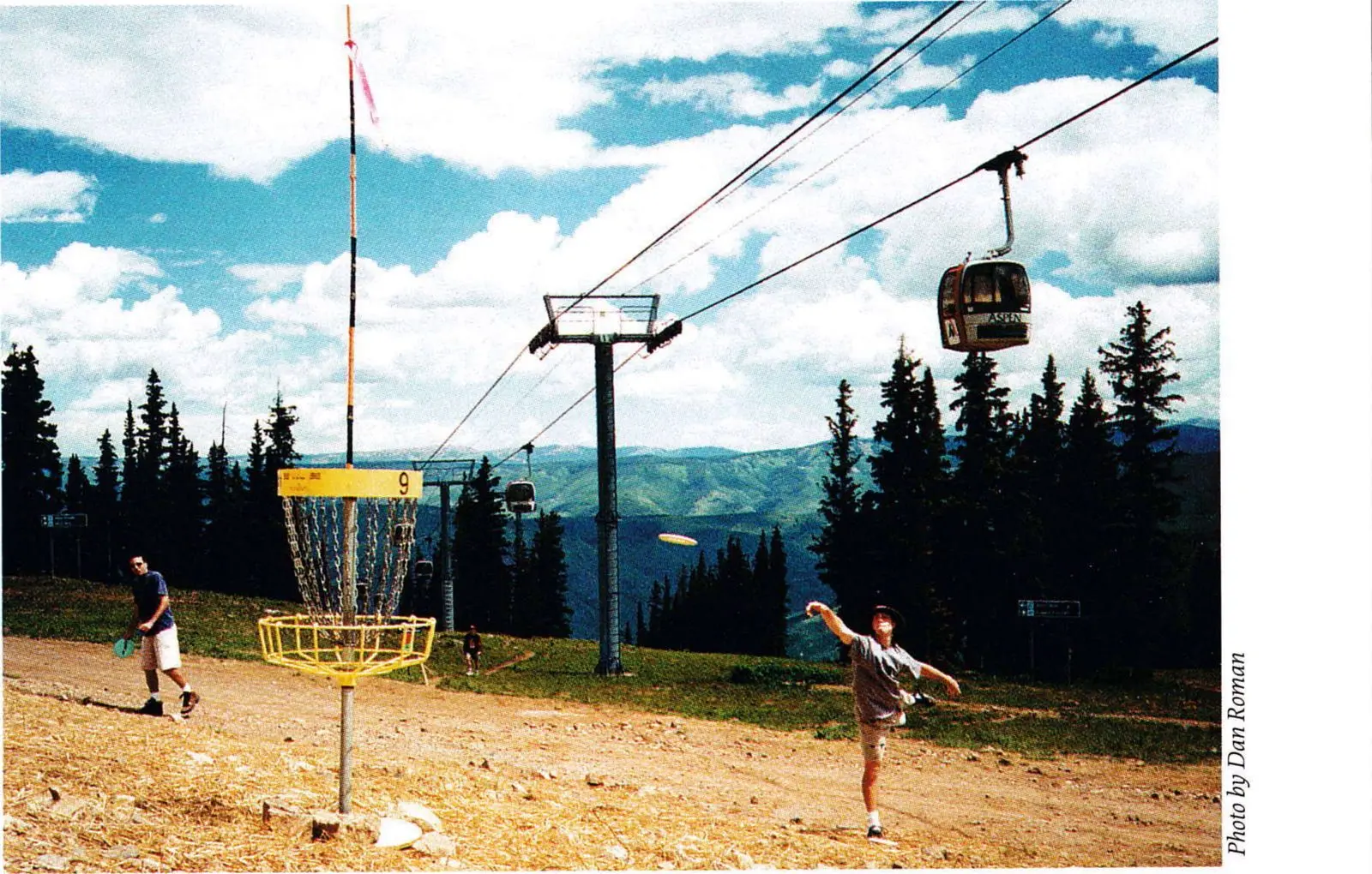 Players throwing discs toward a chain basket on the summit of Ajax Mountain below a gondola line