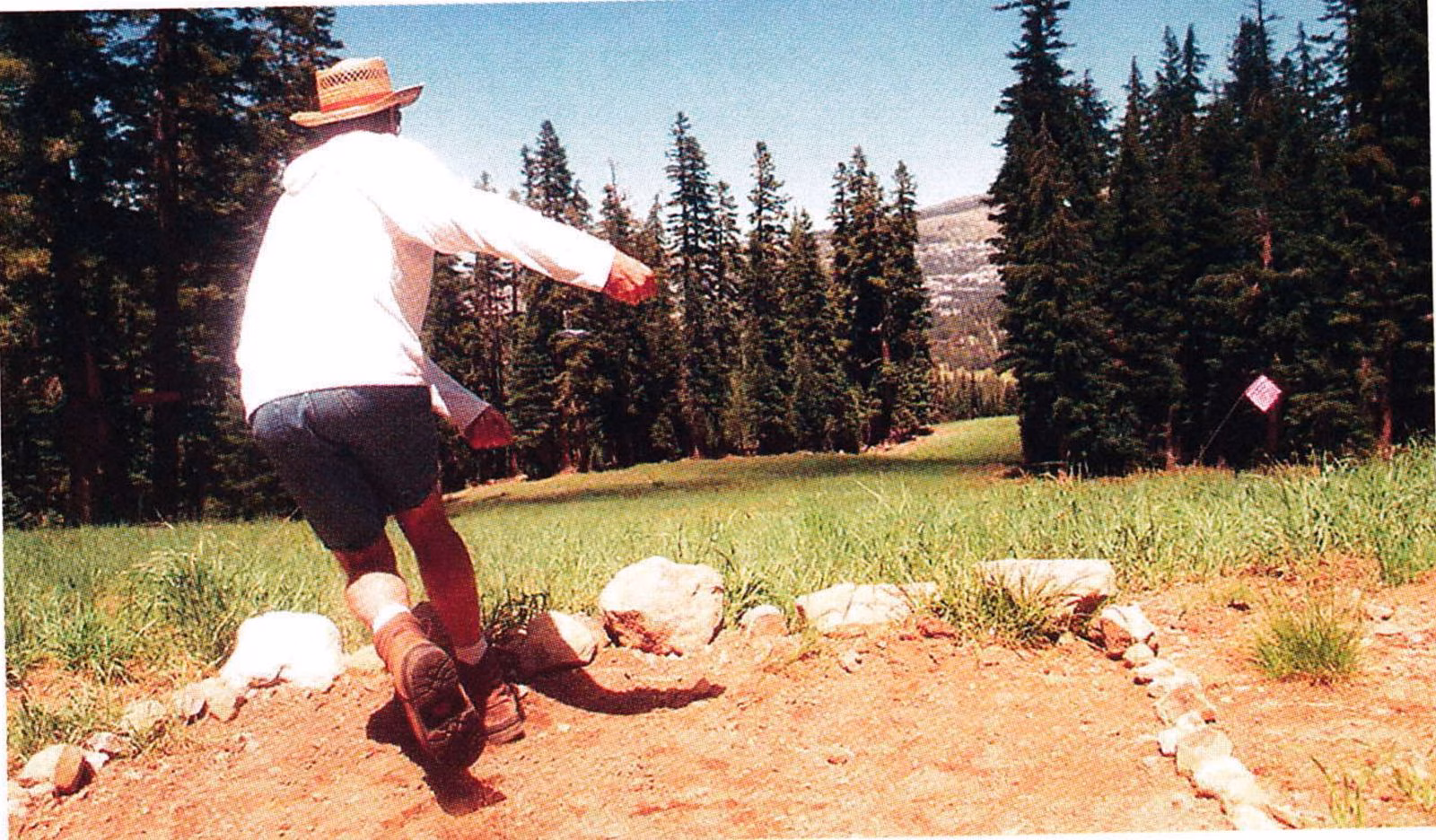 Player in a white shirt and hat throwing a disc from a rock-lined tee in a mountain meadow