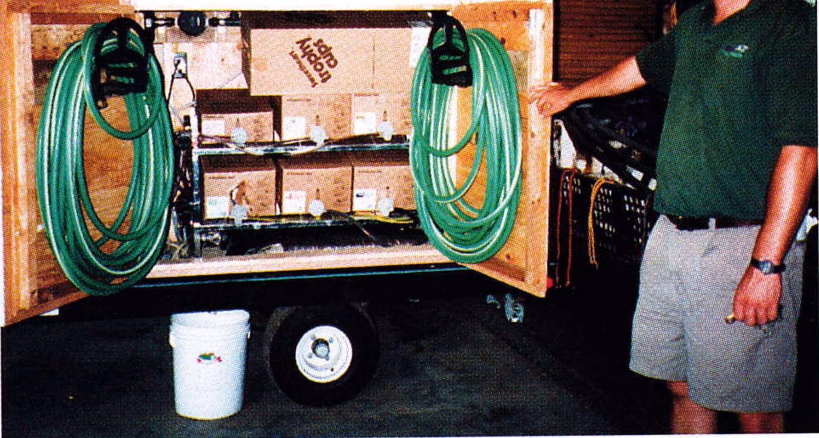 Man standing beside a wooden post-mix soda dispenser cabinet with coiled green hoses mounted on a trailer