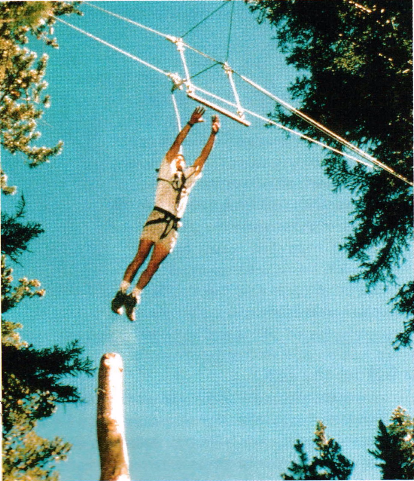 Guest in a harness leaping from a tall pole toward a trapeze against a blue sky