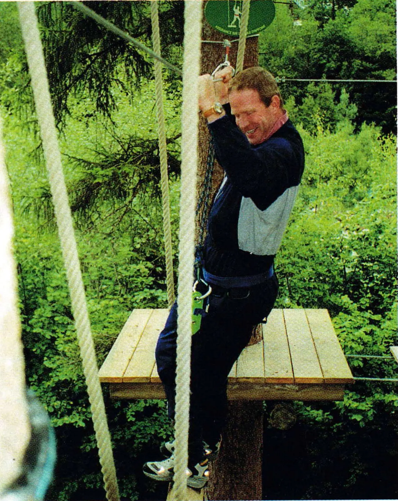 Man in a climbing harness clipped to a cable on a wooden platform among trees