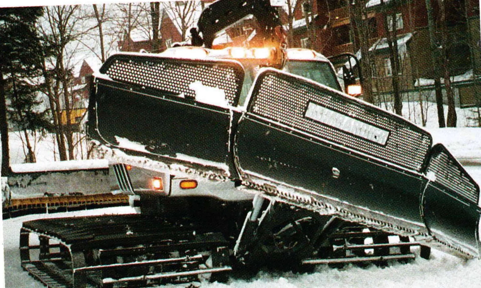 Bombardier groomer with large Terrain Master blade raised, parked in snow near trees
