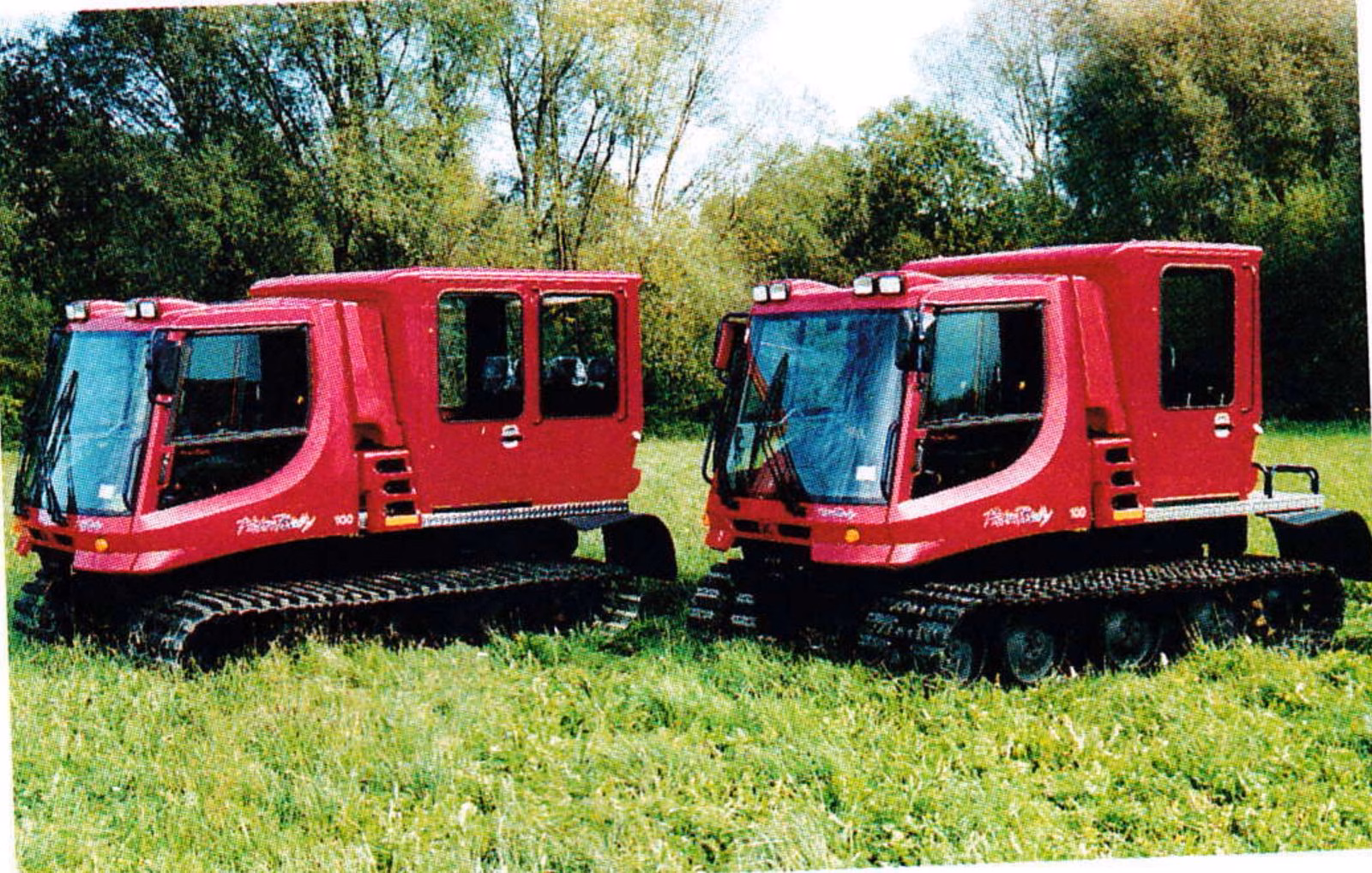 Two red Pisten Bully 100U utility vehicles parked on green grass in front of trees