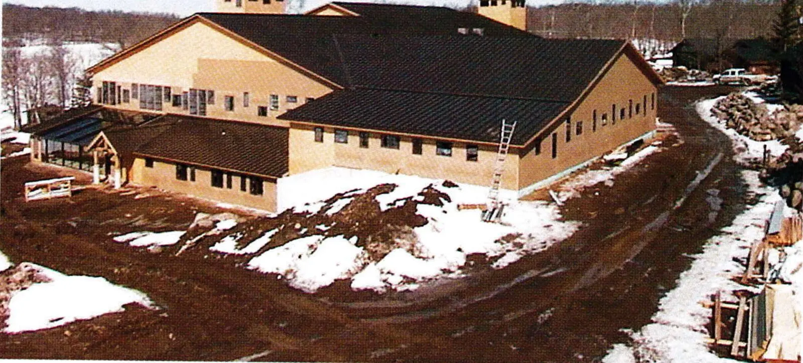 New Maplelag main lodge with brown steel roof and twin lookout towers, seen from above over muddy, partly snow-covered ground